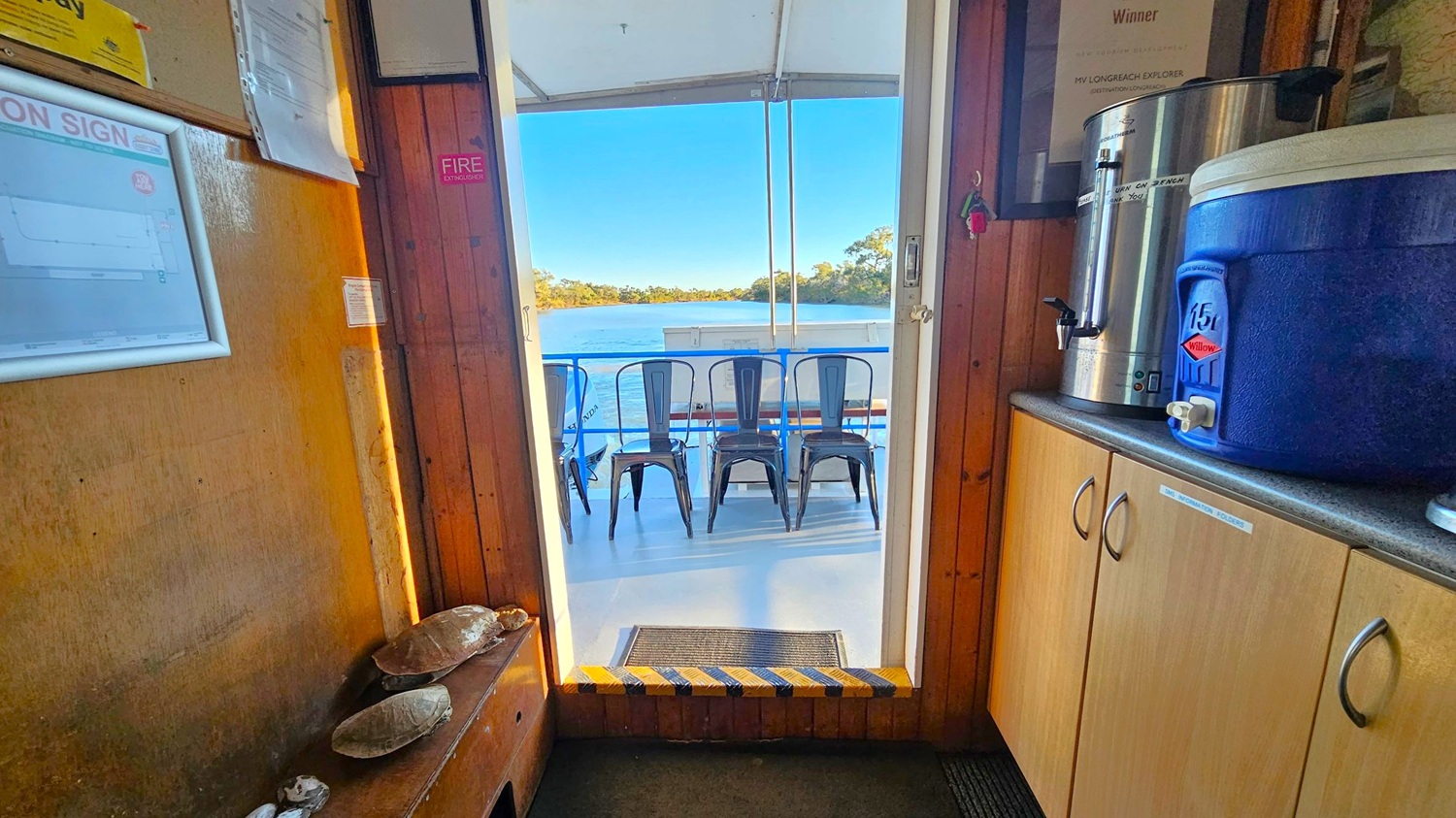 Wooden interior with a notice board on the left wall displaying papers and a fire extinguisher sign. A bench below holds several shells. On the right, a countertop with a large metal urn and a blue water cooler sits atop light-coloured cabinets. Through the open door, a view of a deck with metal chairs facing a body of water and trees in the distance. The floor is carpeted inside and transitions to a smooth surface outside.