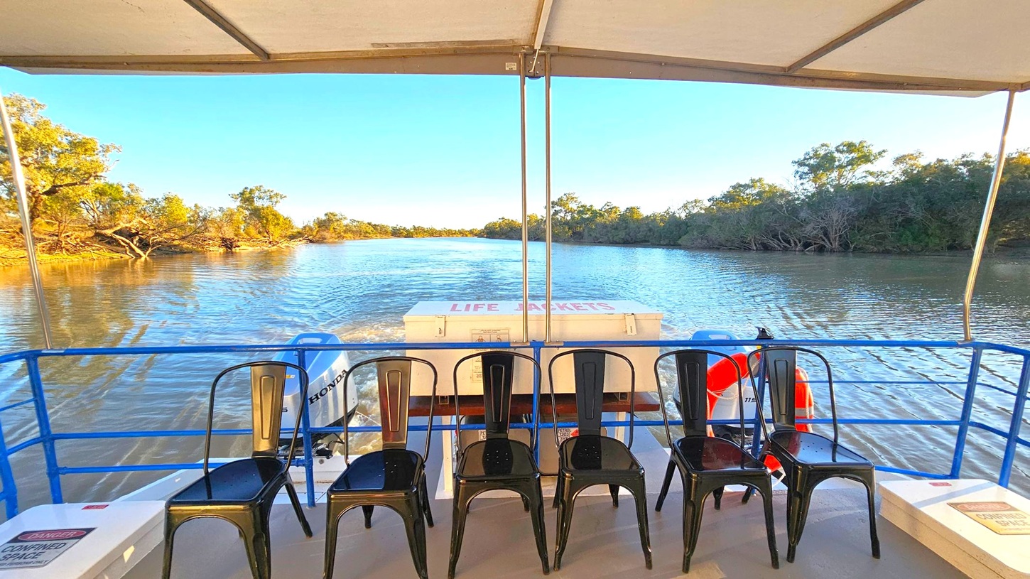 A view from a boat with a canopy overhead, showing five black metal chairs lined up at the back. In the centre, a white box labelled "LIFE JACKETS" is visible. The boat is on a river, with calm water reflecting the clear blue sky. On both sides, there are trees and shrubs along the riverbank. A blue railing runs along the edge of the boat, and a life preserver is attached to it.