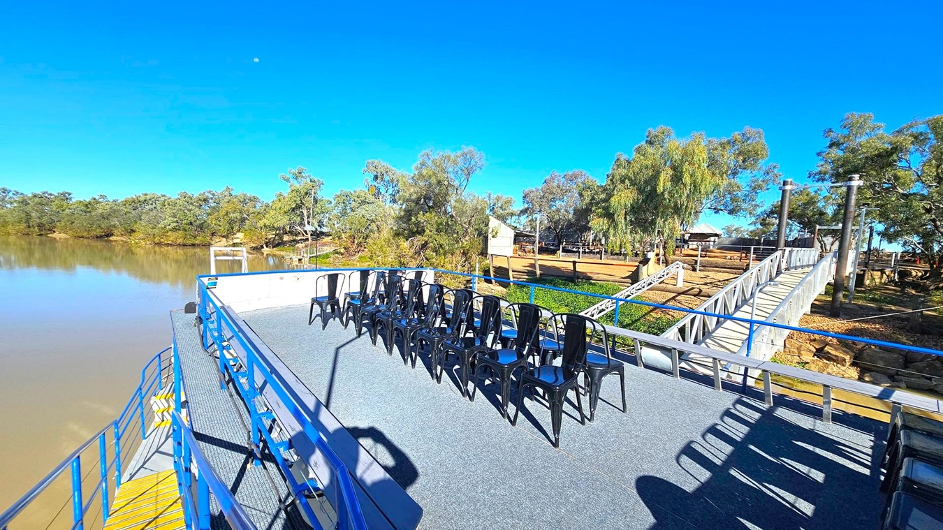 A deck with a blue railing and a grey carpeted floor features a row of black metal chairs. To the right, a metal gangway leads to a grassy area with trees and a small building. The left side shows a calm river bordered by dense trees. The sky is clear and blue.