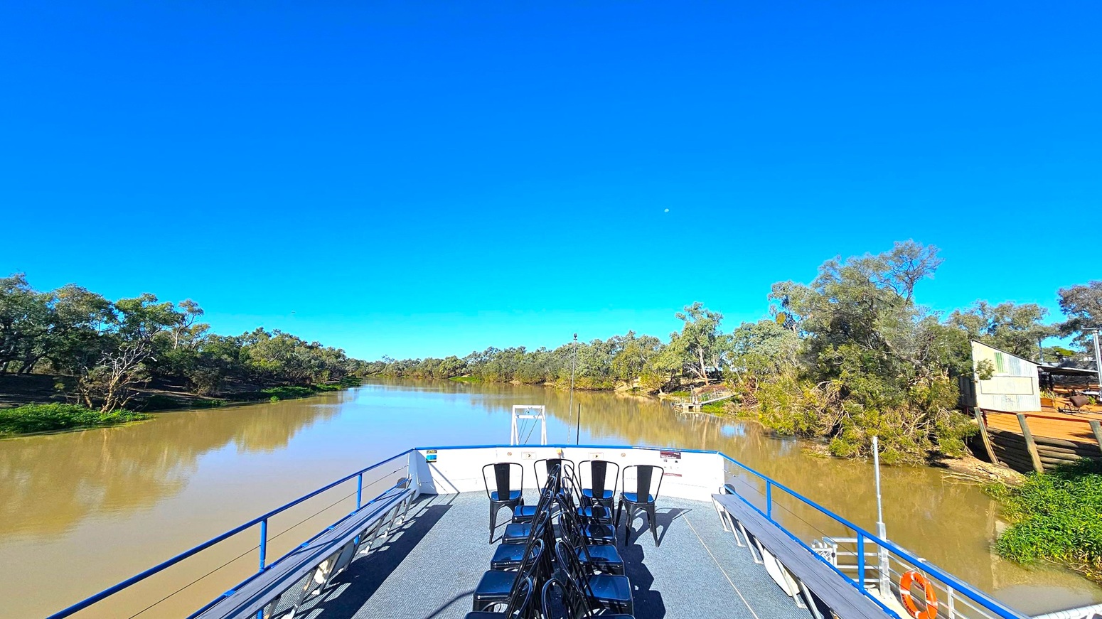 A river scene with a boat deck in the foreground, featuring rows of black chairs. The deck has a blue railing and a grey floor. The river is calm, with muddy water reflecting the surrounding greenery. On the left and right banks, dense trees and shrubs line the river, with a small wooden structure visible on the right. The background shows more trees extending into the distance.