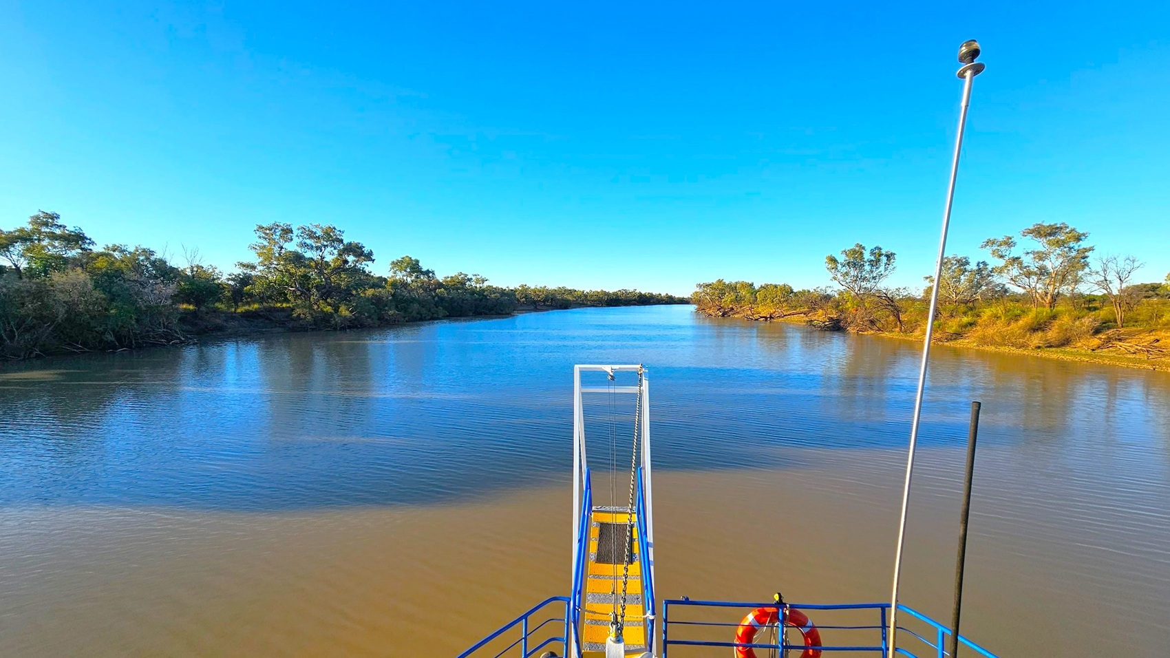 A wide river stretches into the distance, flanked by dense green trees on both sides. In the foreground, a blue and yellow metal structure with a life buoy is visible, possibly part of a boat or dock. A tall metal pole with a device on top extends upwards. The water is calm, reflecting the clear blue sky above. The riverbanks are lined with vegetation, and the scene is tranquil and expansive.