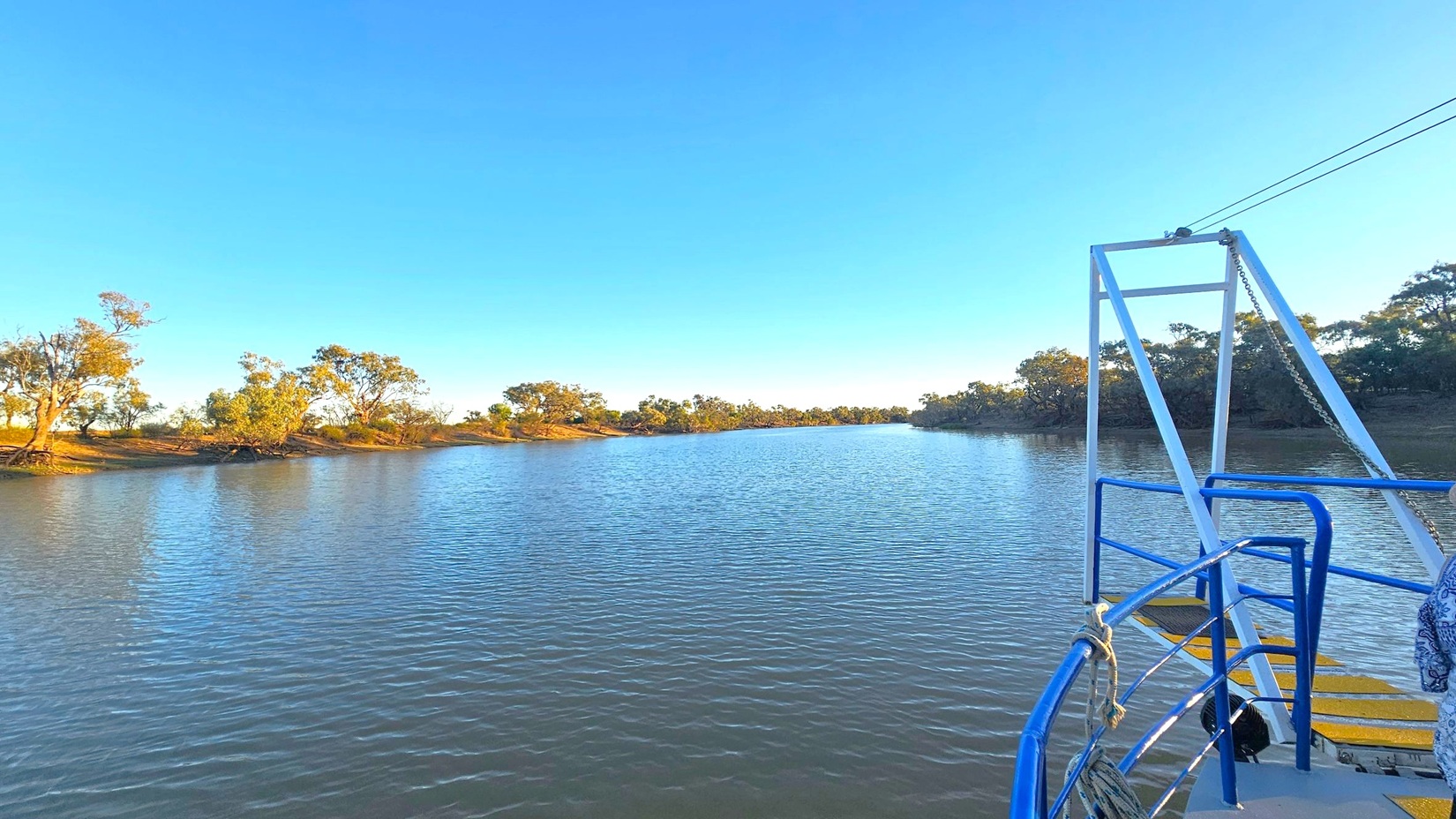 A calm river stretches into the distance, flanked by trees and low vegetation on both sides. The water reflects the surrounding landscape. On the right, part of a boat is visible with blue railings and a white metal structure. A rope is tied to the railing, and a patterned fabric is draped over it. The scene is serene, with the natural environment dominating the view.