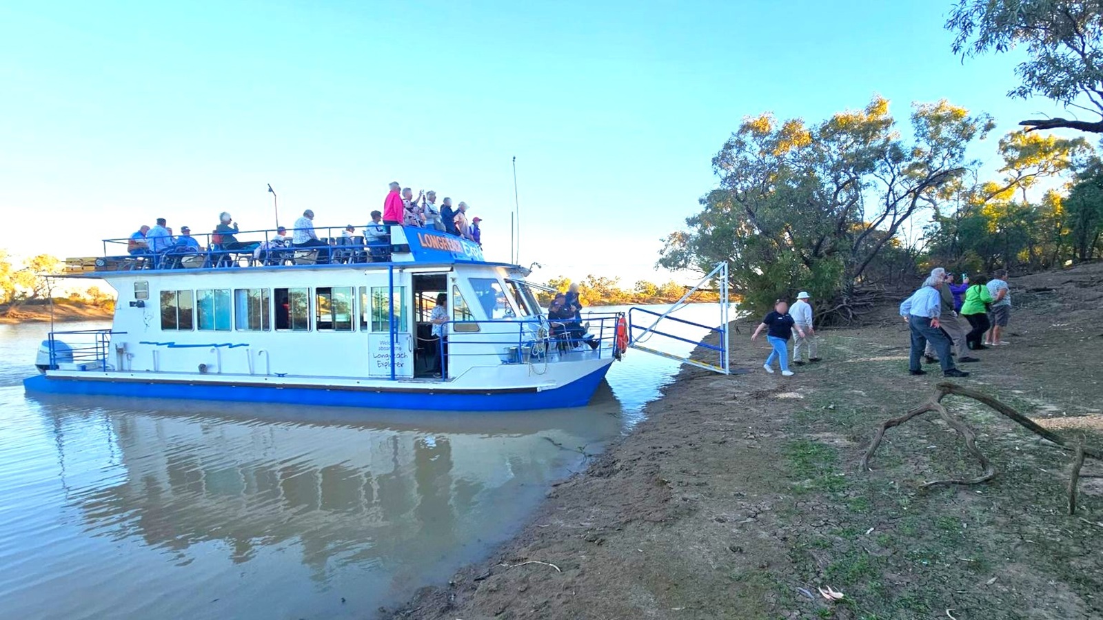 A boat with a blue and white exterior is docked on a riverbank. People are seated on the upper deck, while others are disembarking onto the muddy shore. The riverbank is uneven with patches of green vegetation and scattered branches. Trees with sparse foliage are visible in the background, and the water is calm with gentle ripples. The sky is clear and blue.