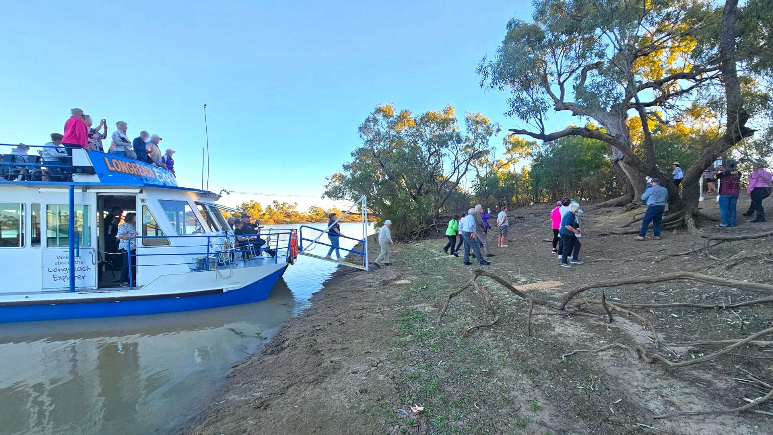 A boat named "Longreach Explorer" is docked on the left, with people standing on its upper deck and others disembarking via a gangway. On the right, a group of people are gathered on a muddy riverbank with scattered greenery. Large trees with twisted branches are in the background, and the scene is set under a clear sky.