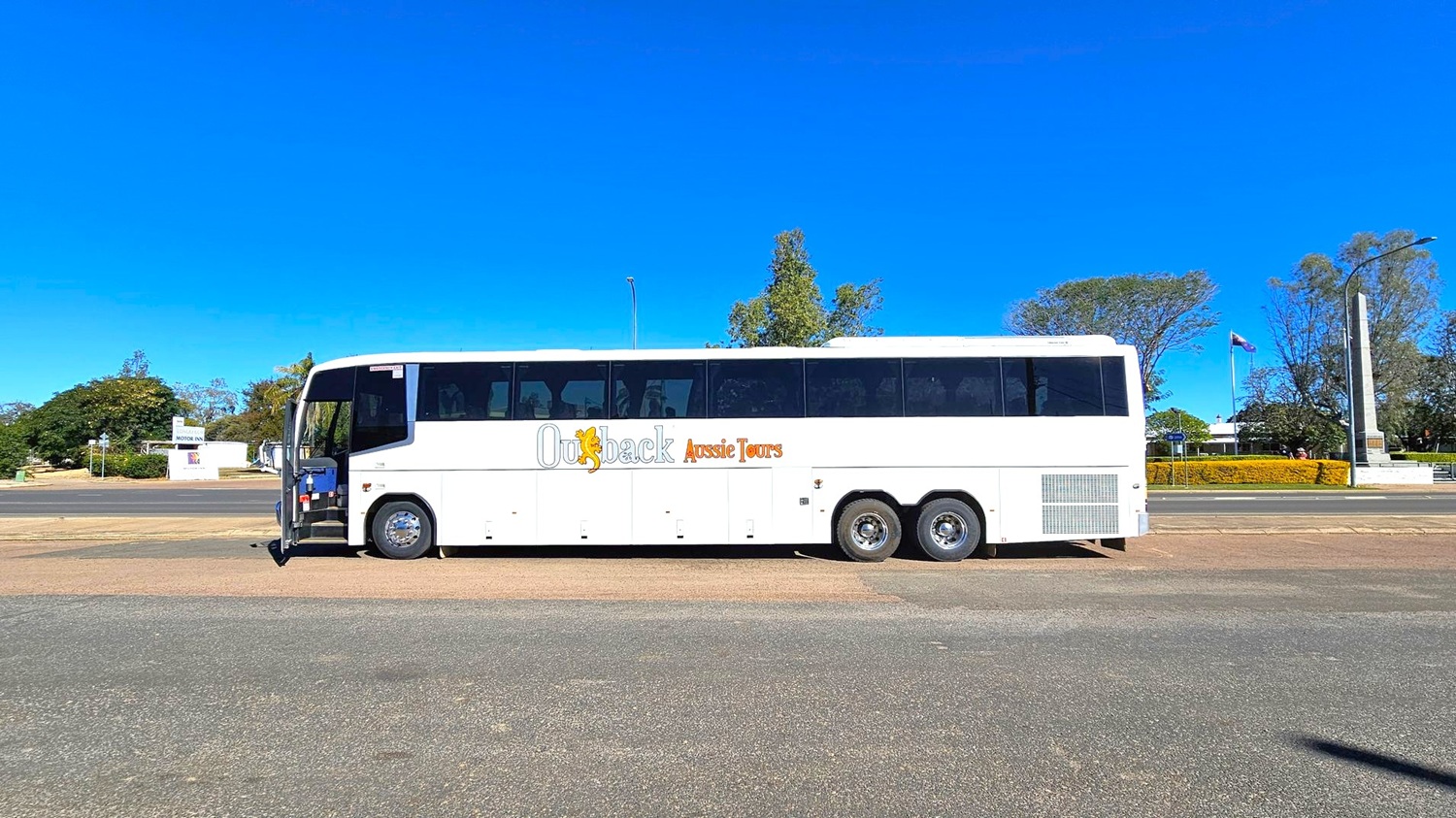 A large white tour bus with "Outback Aussie Tours" written on its side is parked on a paved area. The bus has multiple windows and three axles. In the background, there are trees, a flagpole with a flag, and a tall monument. A sign for a motor inn is visible to the left. The ground is a mix of asphalt and grass, and the sky is clear.