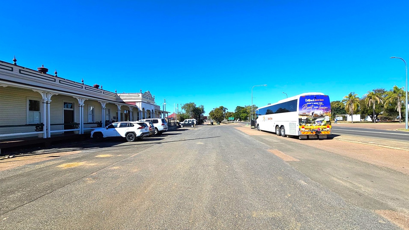 A wide street with a row of parked white cars on the left, adjacent to a long, single-storey building with decorative trim and covered walkways. On the right, a large white coach bus is parked on the road, with palm trees and other greenery in the background. The road surface is a mix of grey and brown patches. The sky is clear and blue.