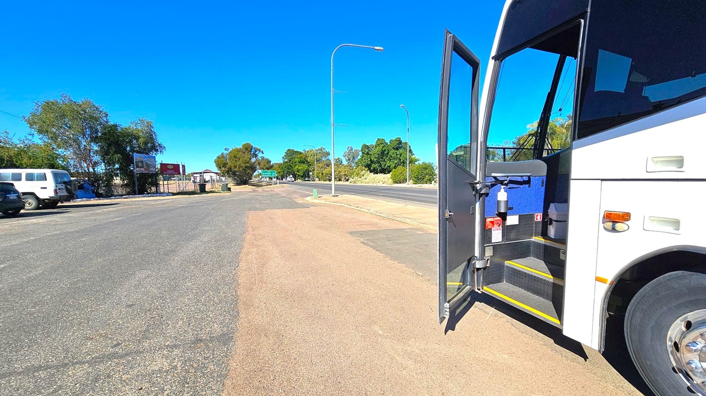 A white bus is parked on the right with its door open, revealing steps and a hand sanitiser dispenser. The road is wide and empty, with a few vehicles parked on the left. Trees and a couple of signboards are visible in the background. The pavement is a mix of asphalt and dirt, and there are streetlights along the road. The scene is well-lit, suggesting daytime.