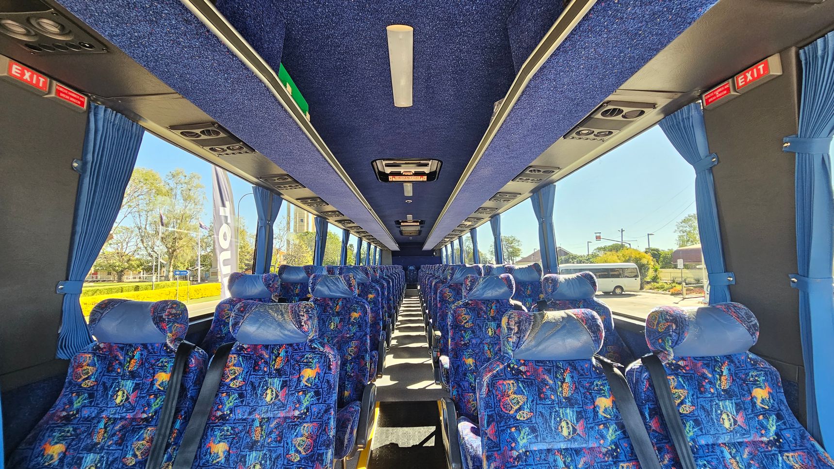 Inside a coach bus, rows of colourful patterned seats with headrests are visible on both sides. Blue curtains hang beside large windows, allowing a view of trees and a parked vehicle outside. The ceiling is lined with blue fabric and features overhead lights and air vents. Red "EXIT" signs are positioned at the front and back. The floor is carpeted in a dark colour, and the aisle runs down the centre.