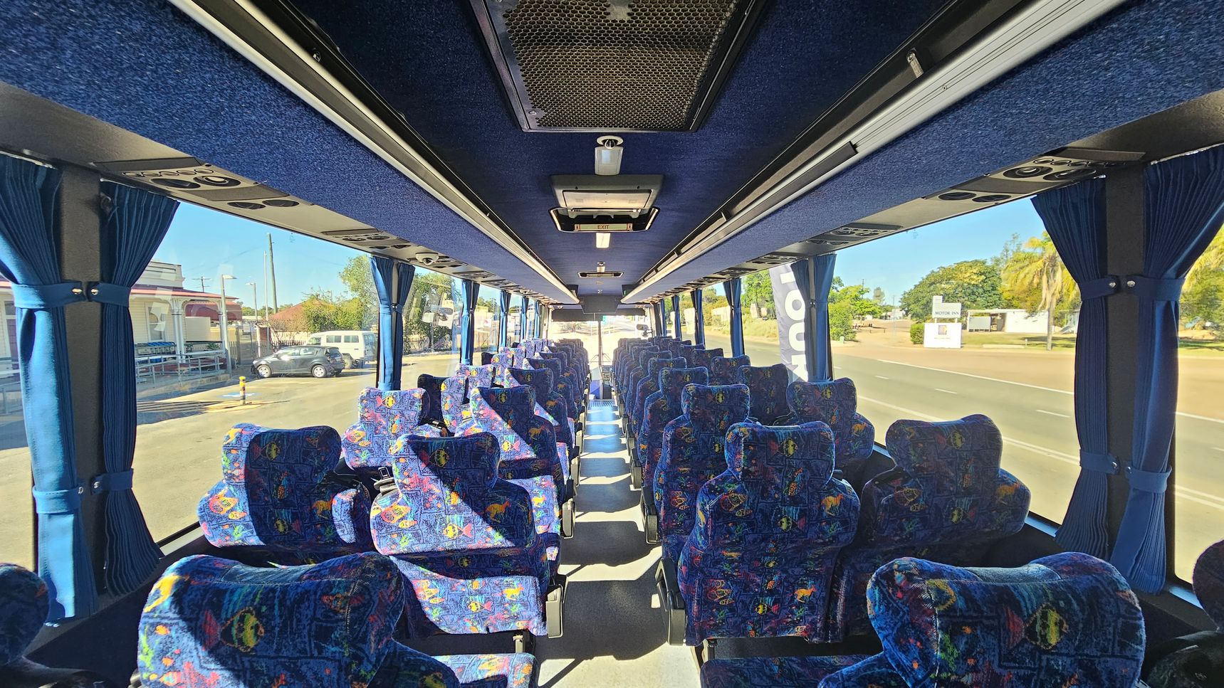 Interior of a coach bus with rows of patterned blue seats on both sides. Blue curtains are drawn back along the large windows. The ceiling is lined with blue fabric and features air vents and lighting. The floor is carpeted in a neutral tone. Outside, a street with parked cars and buildings is visible through the windows.