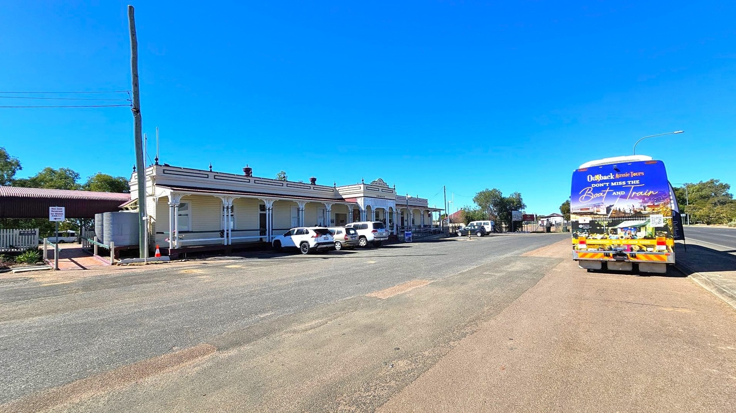 A street scene with a historic building on the left featuring a veranda and several parked white vehicles. A large water tank and a tall pole are visible nearby. On the right, a colourful tour bus with promotional graphics is parked on the road. The road is wide and mostly empty, with a few trees and a small building in the background. The sky is clear and blue.