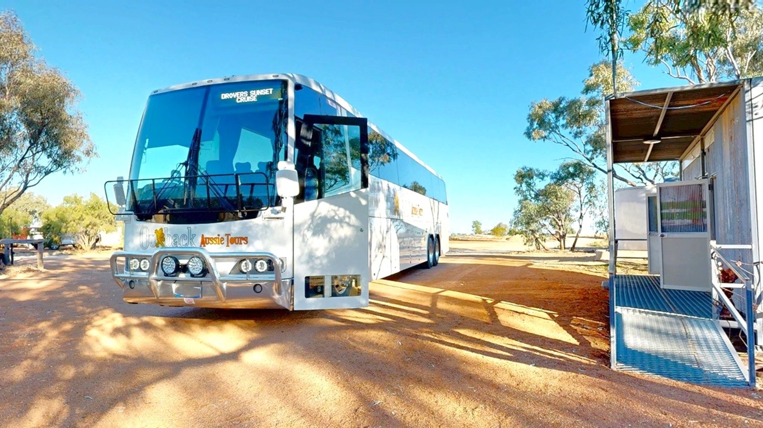 A large white tour bus is parked on a dirt road, with its front door open. The bus has a sign indicating a sunset tour. To the right, there is a small metal structure with a ramp and railing. Trees with sparse foliage are scattered around, casting shadows on the ground. The sky is clear and blue.