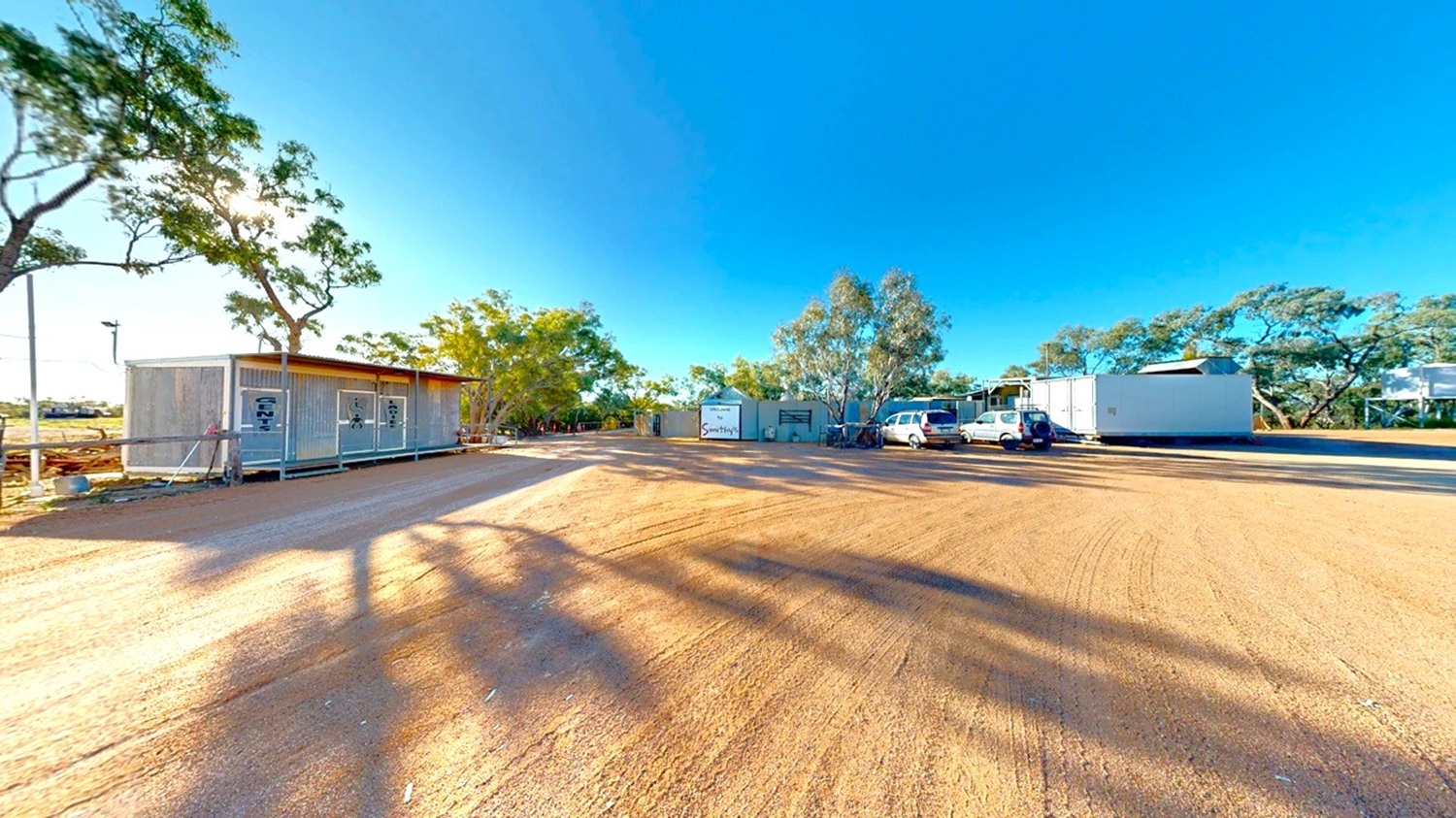 A wide, open area with a dirt ground. On the left, there is a small, rectangular building with a metal exterior and a flat roof, surrounded by a few trees. On the right, several vehicles are parked near another building, which is larger and also has a metal exterior. In the background, more trees are visible, casting long shadows across the ground. The sky is clear and blue.