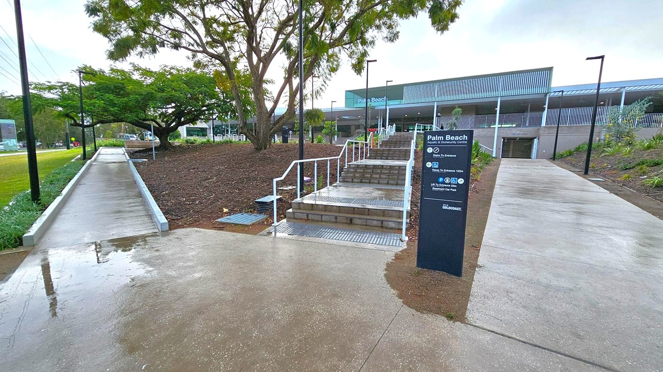 A concrete pathway leads to a set of stairs with white railings in the centre, flanked by a sign for Palm Beach Aquatic & Community Centre. To the left, a sloped path runs alongside a grassy area with a large tree. On the right, another path leads to the building entrance. The building in the background has a modern design with a large overhang and louvred facade. The ground is wet, suggesting recent rain, and there are street lamps along the paths.