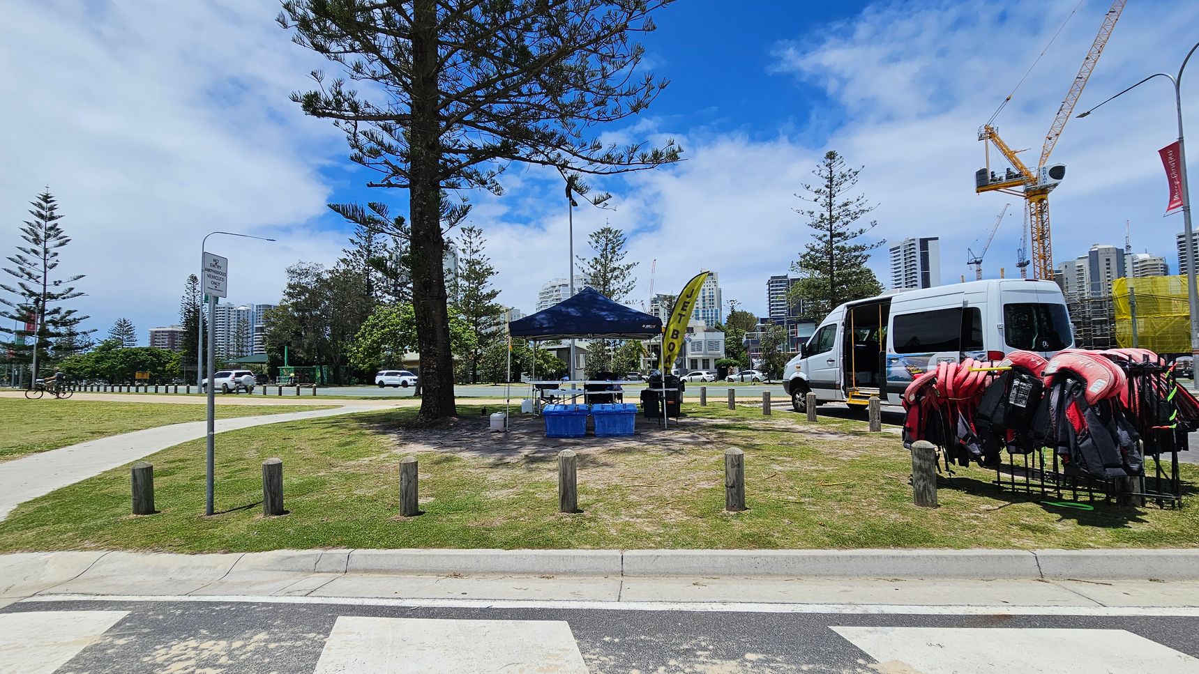 A park area with a small blue tent in the centre, surrounded by trees. To the right, a white van is parked with its door open, and a rack of life jackets is visible. A yellow flag with text is near the tent. In the background, there are tall buildings and a construction crane. A cyclist is on the left, and a signpost is visible. The ground is grassy with a paved path, and wooden bollards line the area.