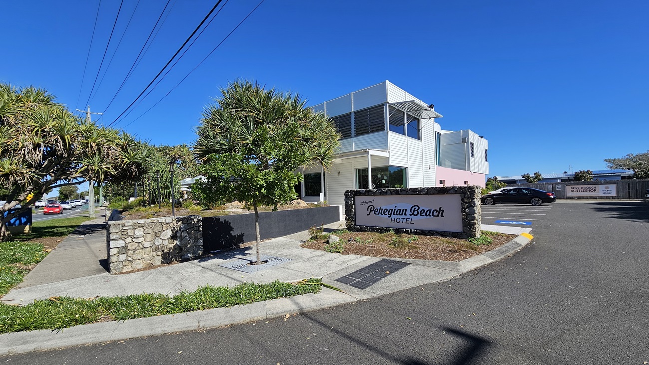 On the left, there is a stone wall with a tree and a pathway leading to the entrance of the Peregian Beach Hotel. The hotel sign is in front of the stone wall. In the middle, the hotel building has a modern design with large windows and a balcony on the upper floor. On the right, there is a car park with several vehicles. The foreground has a paved road lined with trees and power lines. The background includes more trees and parts of the hotel building.