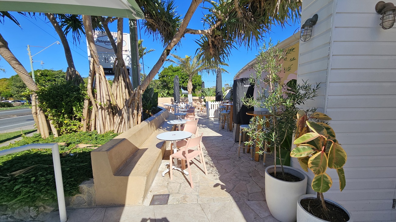 On the left, there is a beige bench along a stone wall with a tree and plants. In the middle, there are several tables and chairs arranged on a stone floor, with umbrellas providing shade. On the right, there is a white wooden wall with potted plants placed along it. The background includes more trees and a glimpse of the road with a visible hotel sign. The ceiling is formed by the tree canopy providing partial shade.
