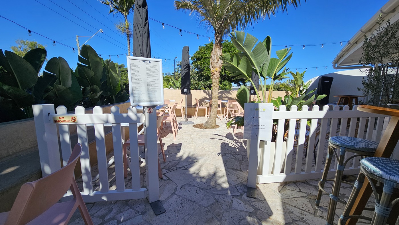 On the left, there are pink chairs and tables next to a white picket fence, with large green plants behind them. In the middle, there is a stone floor with more pink chairs and tables under black umbrellas, surrounded by palm trees and string lights overhead. On the right, there is a white building with a bar counter and stools. The background includes more green plants and a beige wall. The ceiling consists of string lights hanging above the seating area.