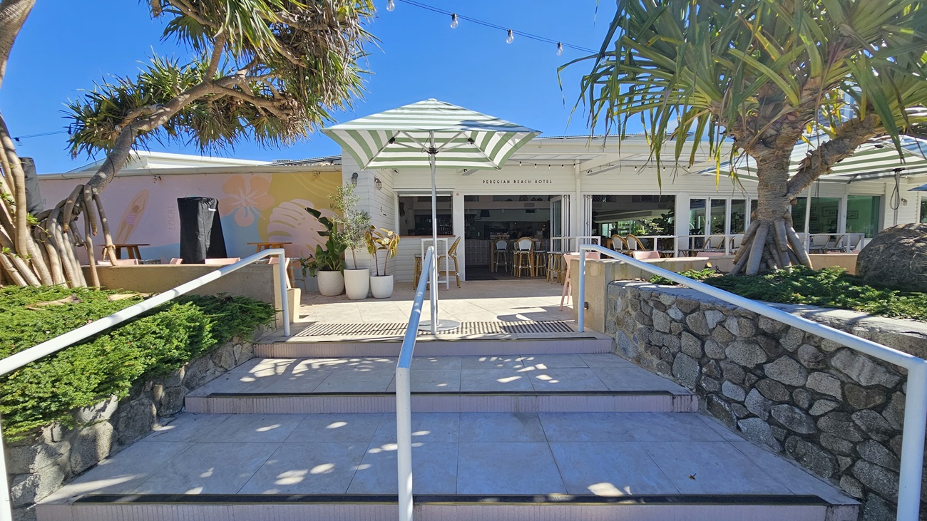 On the left, there are large green plants and trees next to a stone wall. In the middle, there are steps leading up to the entrance of the Peregian Beach Hotel, with white railings and potted plants. The entrance has a green and white striped umbrella and a few tables and chairs. On the right, there are more large green plants and trees next to another stone wall. The floor is tiled, and the ceiling has string lights hanging above the entrance area.