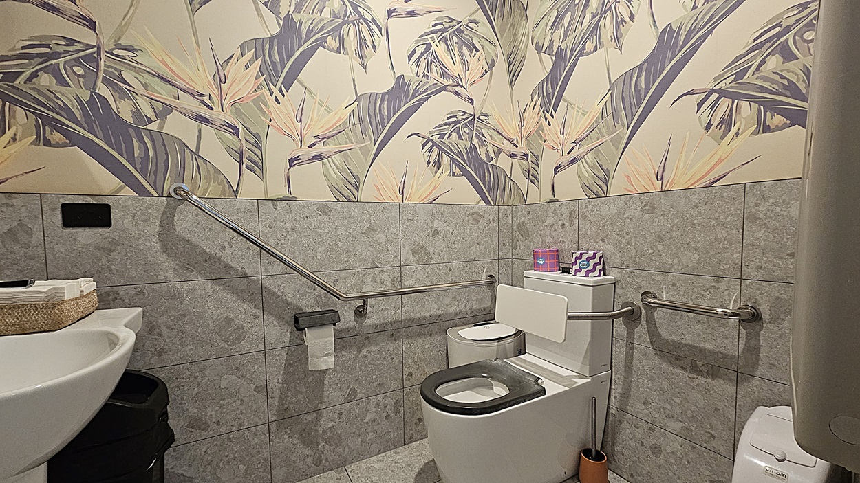 On the left, there is a white door with a wheelchair access sign and a door handle, followed by a wall-mounted baby changing station. On the right, there is a white panelled wall with a black ATM machine. In the middle, there is a long, narrow corridor with a polished concrete floor and white walls. The background features another white door at the end of the corridor. The ceiling is white with recessed lighting and a smoke detector.
