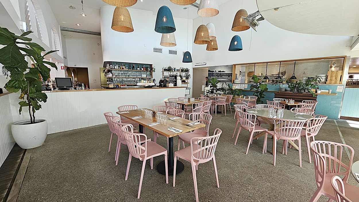 On the left, there is a white counter with a geometric pattern and a potted plant on top. In the middle, there are round tables with pink chairs on a grey carpeted floor, under hanging wicker and blue lamps. The background features a turquoise counter with plants and shelves, and a staircase leading to an upper level. On the right, there is a menu stand on a gold pole, more plants, and a wooden floor section. The ceiling is white with recessed lighting and hanging lamps.
