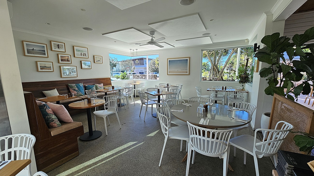 On the left, there are wooden booths with colourful cushions and round tables, with framed pictures on the wall above. In the middle, there are round tables with white chairs on a speckled floor, leading to large windows that provide a view of trees and outdoor seating. On the right, there are more white chairs and tables near a large window with a potted plant beside it. The ceiling is white with recessed lighting and ceiling fans.