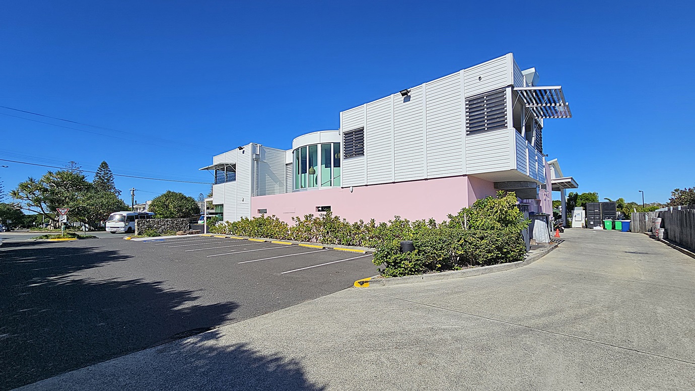 On the left, there is a white building with large windows and a pink lower section, surrounded by landscaped greenery. There are a few parked cars along the side. On the right, there is a driveway leading to a parking area with green, yellow, and blue bins. In the middle, there is an asphalt parking lot with marked spaces and a concrete walkway. The background includes more buildings and trees.