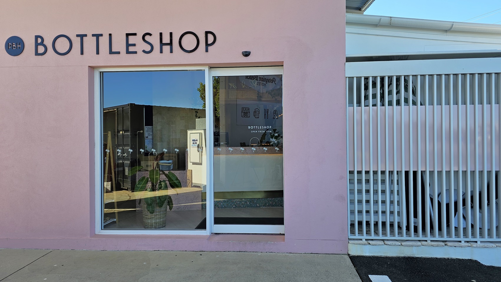 On the left, there is a pink wall with a large window and a glass door, both displaying the word "BOTTLESHOP" above them. Inside the window, there are shelves with bottles and a potted plant. On the right, there is a white slatted fence with a staircase behind it. The floor is a mix of concrete and asphalt. The ceiling is not visible.