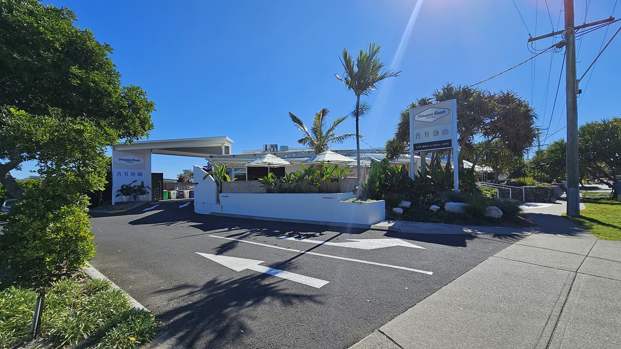 On the left, there is a large tree and a landscaped area with green bushes. On the right, there is a concrete pavement with a tall utility pole and a grassy area. In the middle, there is an entrance to a building with a white canopy, surrounded by palm trees and other plants. There are two signs with the text "Peregian Beach Hotel" near the entrance. The floor is asphalt with white directional arrow.