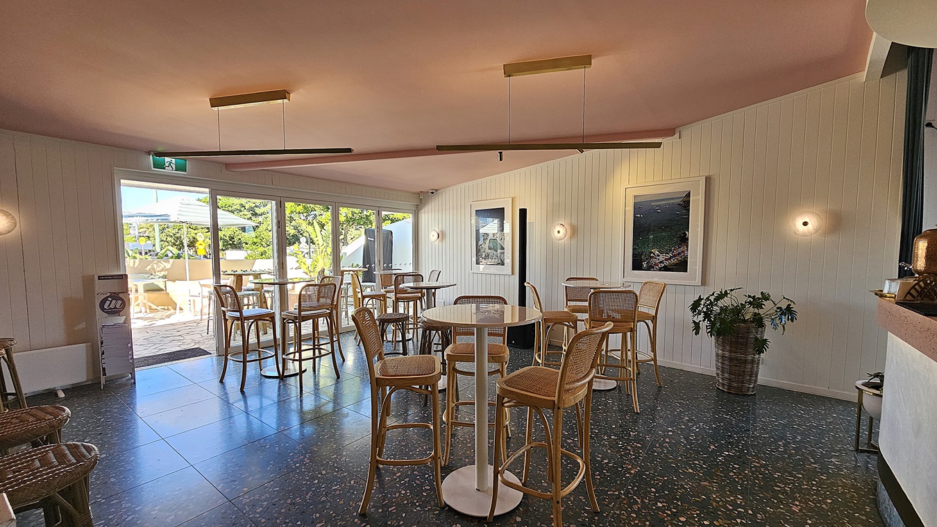 On the left, there are tall wicker stools around high tables near large glass doors that open to an outdoor seating area. In the middle, there are round tables with wicker chairs on a speckled floor. On the right, there is a white wall with framed pictures and a potted plant. The ceiling is pink with recessed lighting and long, rectangular hanging lights.