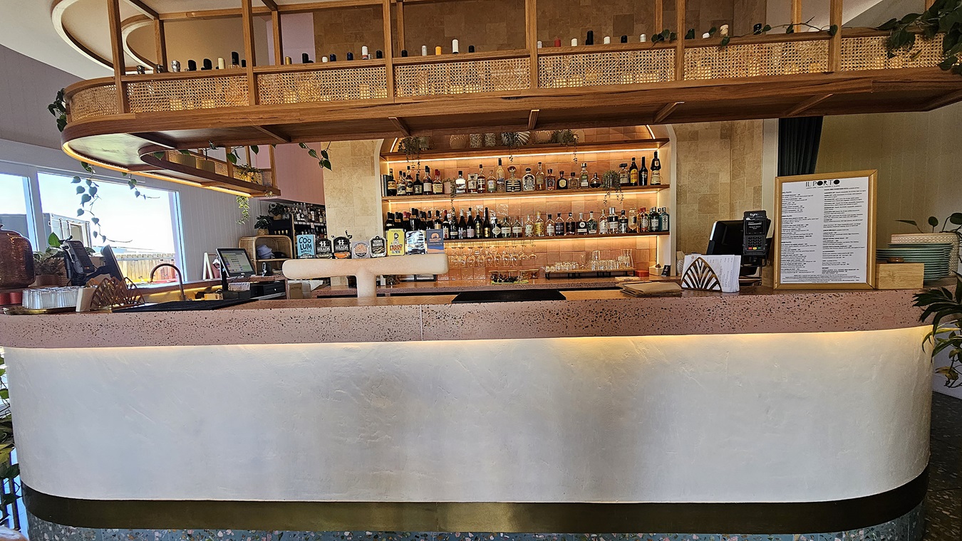 On the left, there are plants and decorative items on a counter, with a glimpse of outdoor seating through a window. In the middle, there is a curved bar counter with a pink terrazzo top and a white front, displaying various bottles and a menu. The background features shelves filled with bottles and glasses, with a wooden wall behind them. On the right, there is a menu stand and more plants. The floor is speckled, and the ceiling has wooden panels with recessed lighting and hanging shelves.