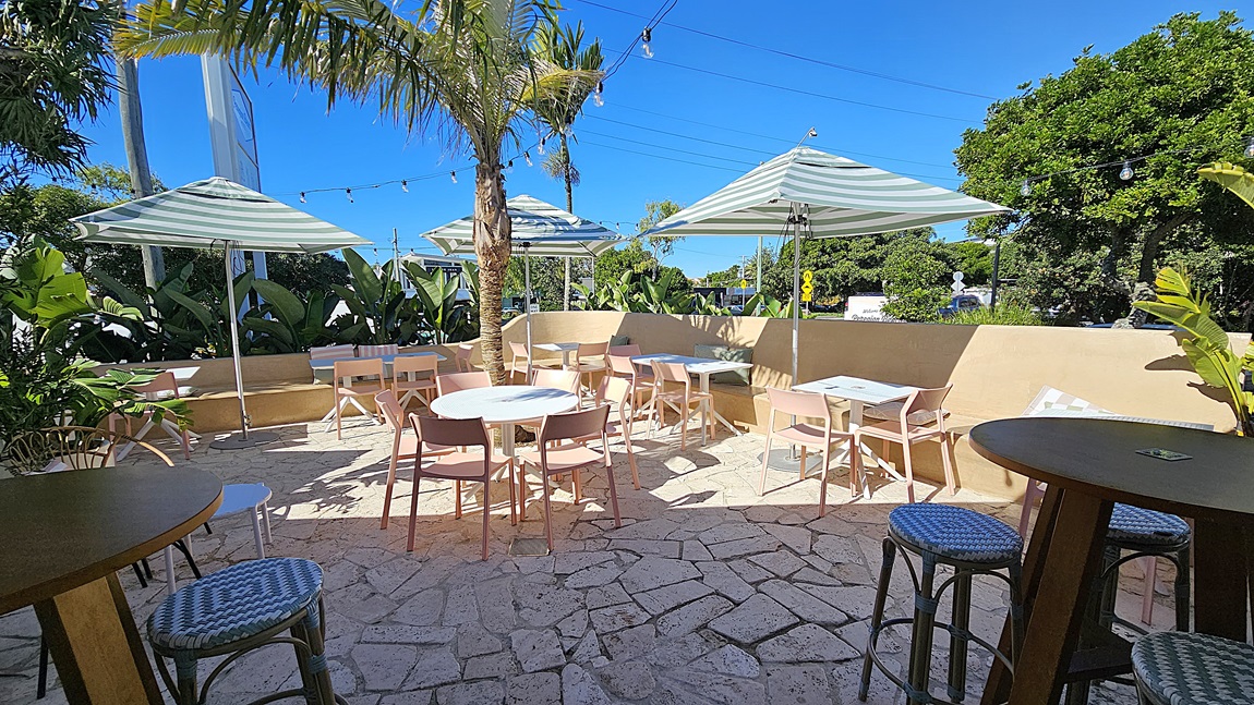 On the left, there are round wooden tables with wicker stools, partially shaded by a large striped umbrella. In the middle, there are several white tables with pink chairs arranged on a stone patio, centred around a palm tree. The background includes more tables and chairs, surrounded by lush green plants and additional striped umbrellas. On the right, there are more wooden tables and wicker stools, with string lights hanging above. The floor is made of irregular stone tiles.