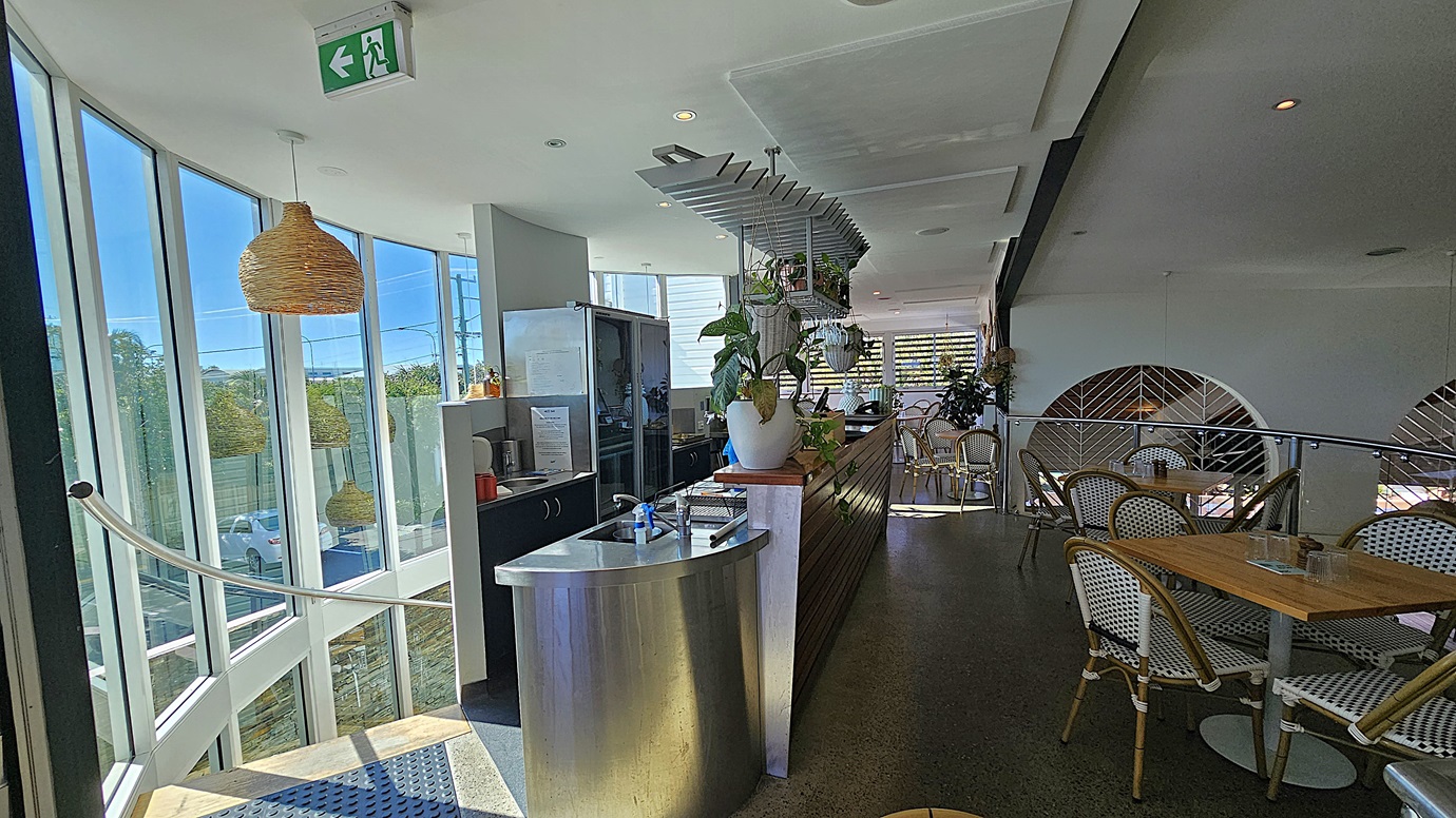 On the left, there is a curved staircase with metal railings leading down, next to large windows. In the middle, there is a stainless steel counter with a potted plant and a coffee machine, with hanging shelves above. The background features more windows and a view of trees outside. On the right, there are wooden tables with wicker chairs, and a partition with a geometric design. The floor is polished concrete, and the ceiling is white with recessed lighting and a hanging wicker lamp.