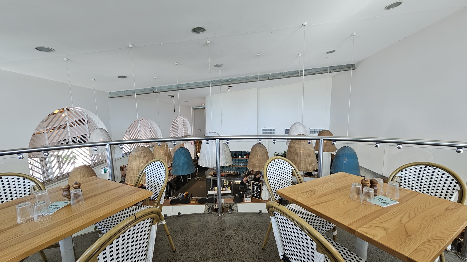 On the left, there are wooden tables with white and blue woven chairs, set with glasses and condiments. In the middle, there is a glass railing overlooking the lower level, with hanging wicker and metal lamps of various shapes and sizes. The background features a white wall with a ventilation duct near the ceiling. On the right, there are more tables and chairs similar to those on the left. The floor is polished concrete, and the ceiling is white with recessed lighting and hanging lamps.