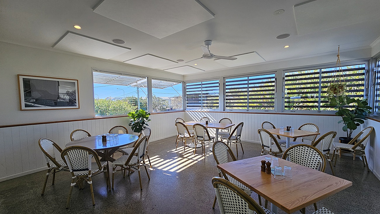 On the left, there are wooden tables with white and green woven chairs, next to a white wall. In the middle, there are more tables and chairs on a polished concrete floor, with sunlight streaming through louvred windows. The background features large windows with louvres and a view of greenery outside. On the right, there are additional tables and chairs, along with potted plants near the windows. The ceiling is white with recessed lighting, ceiling fans, and acoustic panels.