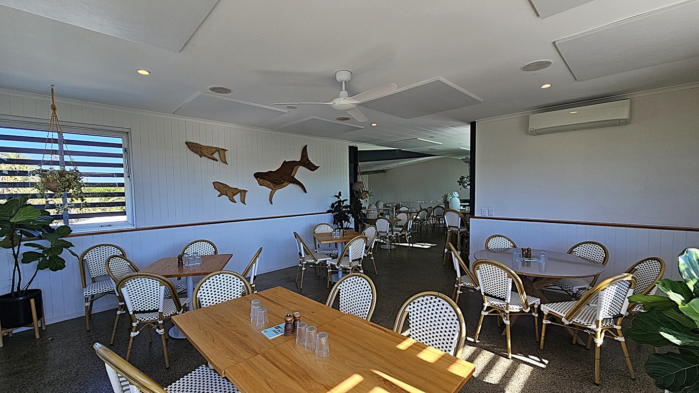 On the left, there are wooden tables with white and green woven chairs, a potted plant, and a window with horizontal louvres. On the right, there are more tables and chairs, a potted plant, and a white wall with an air conditioning unit. In the middle, there are additional tables and chairs on a dark polished floor, with a wall featuring wooden fish decorations. The background has more seating and a large mirror. The ceiling is white with recessed lighting, a ceiling fan, and acoustic panels.