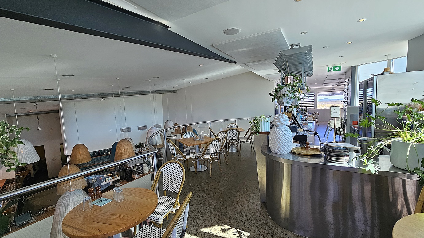 On the left, there are round wooden tables with white and gold woven chairs, and hanging wicker lamps. On the right, there is a curved metal counter with potted plants, plates, and a large white pineapple decoration. In the middle, there are more tables and chairs on a polished concrete floor, with a glass railing overlooking the lower level. The background features large windows with louvres and a view of a distant landscape. The ceiling is white with recessed lighting and exposed beams.