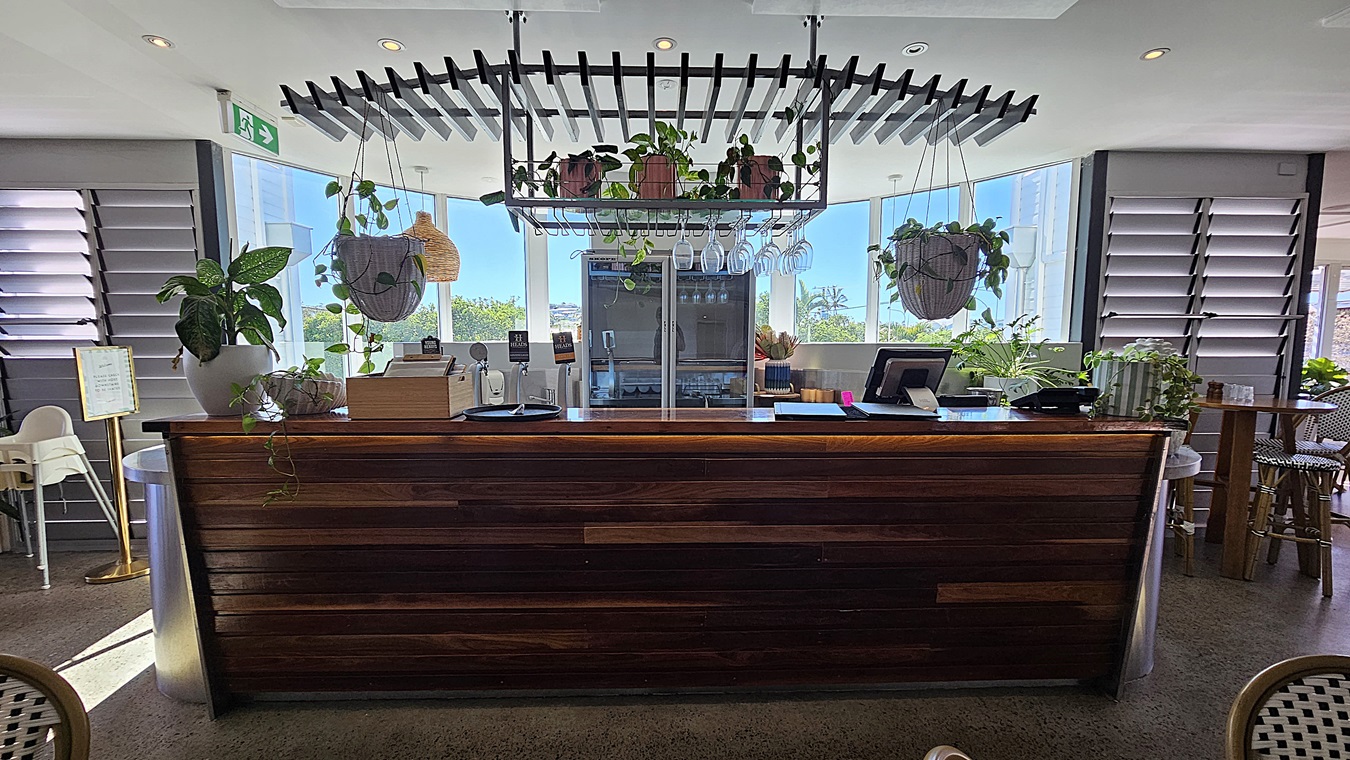 On the left, there are louvred windows with potted plants on the windowsill and a white high chair. On the right, there are more louvred windows, a small wooden table with chairs, and additional potted plants. In the middle, there is a dark wooden counter with a metal top, hanging plants, and glasses above it. The floor is polished concrete, and the ceiling is white with recessed lighting and a black slatted structure above the counter.