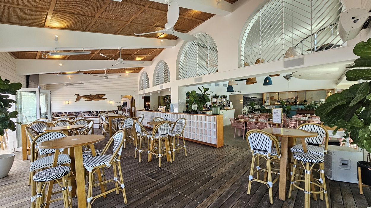 On the left, there are round wooden tables with wicker chairs featuring black and white patterned cushions, set on a wooden floor. In the middle, there is a bar counter with a white front and high stools, with plants and decor on top. The background includes arched windows with decorative patterns and more seating. On the right, there are additional tables and wicker chairs, with a mix of plants and hanging lights. The ceiling has wooden panels, white beams, ceiling fans, and recessed lighting.