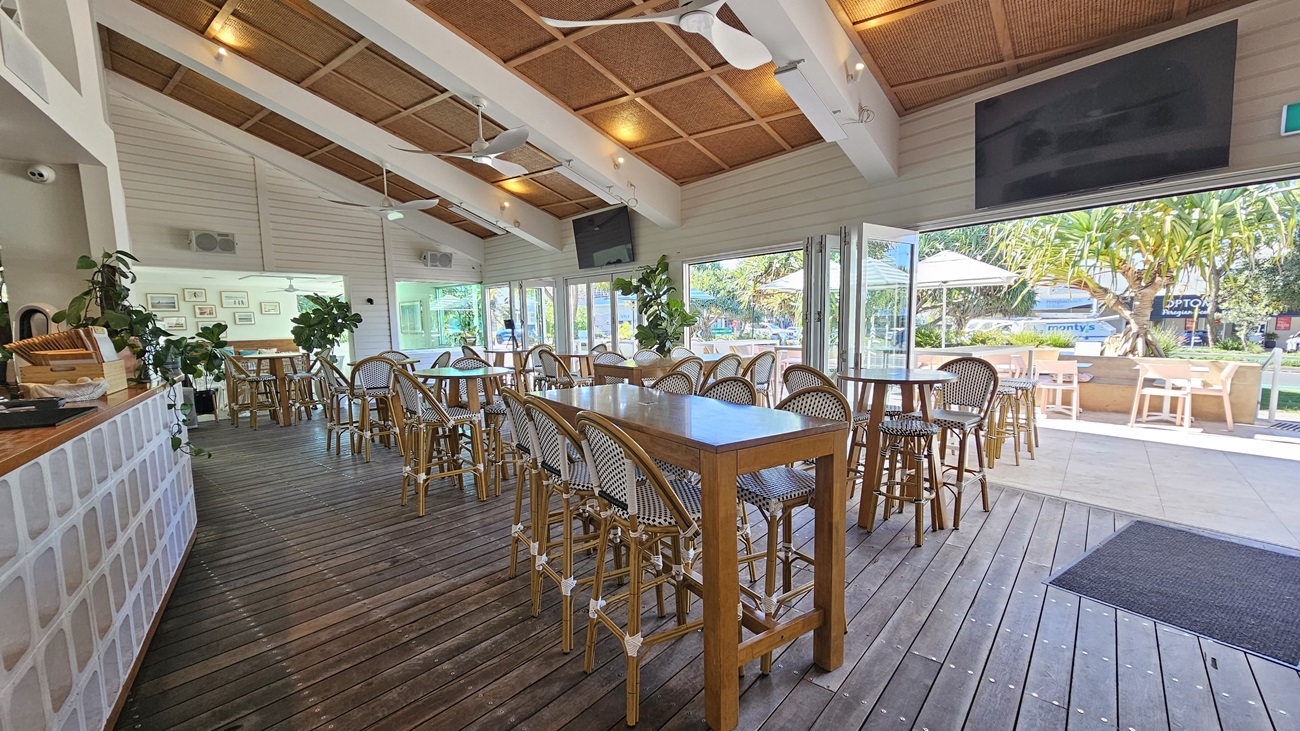 On the left, there is a counter with a white brick front and a wooden top, with plants and a coffee machine. In the middle, there are wooden tables and wicker chairs on a wooden floor, with large glass sliding doors leading to an outdoor seating area. On the right, there are more tables and chairs on a tiled floor, with large windows showing greenery and outdoor umbrellas. The ceiling has white beams, wooden panels, ceiling fans, and recessed lighting.