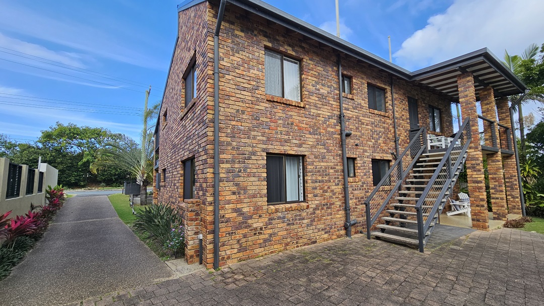 On the left, there is a paved driveway lined with red-leaved plants and a white fence. In the middle, there is a two-storey brick house with multiple windows and an external staircase leading to the upper floor. On the right, there is a small patio area with white chairs and a table, surrounded by tall plants. The floor is a mix of paving stones and asphalt.