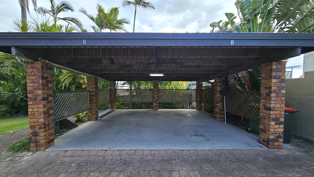 On the left, there is a brick pillar supporting the carport roof, with a lattice fence and lush green plants behind it. In the middle, there is an open concrete floor space under the carport, with a light fixture on the wooden ceiling. On the right, there is another brick pillar, a lattice fence, and more green plants. The floor is a mix of concrete under the carport and paving stones in the foreground. The ceiling is wooden with exposed beams.