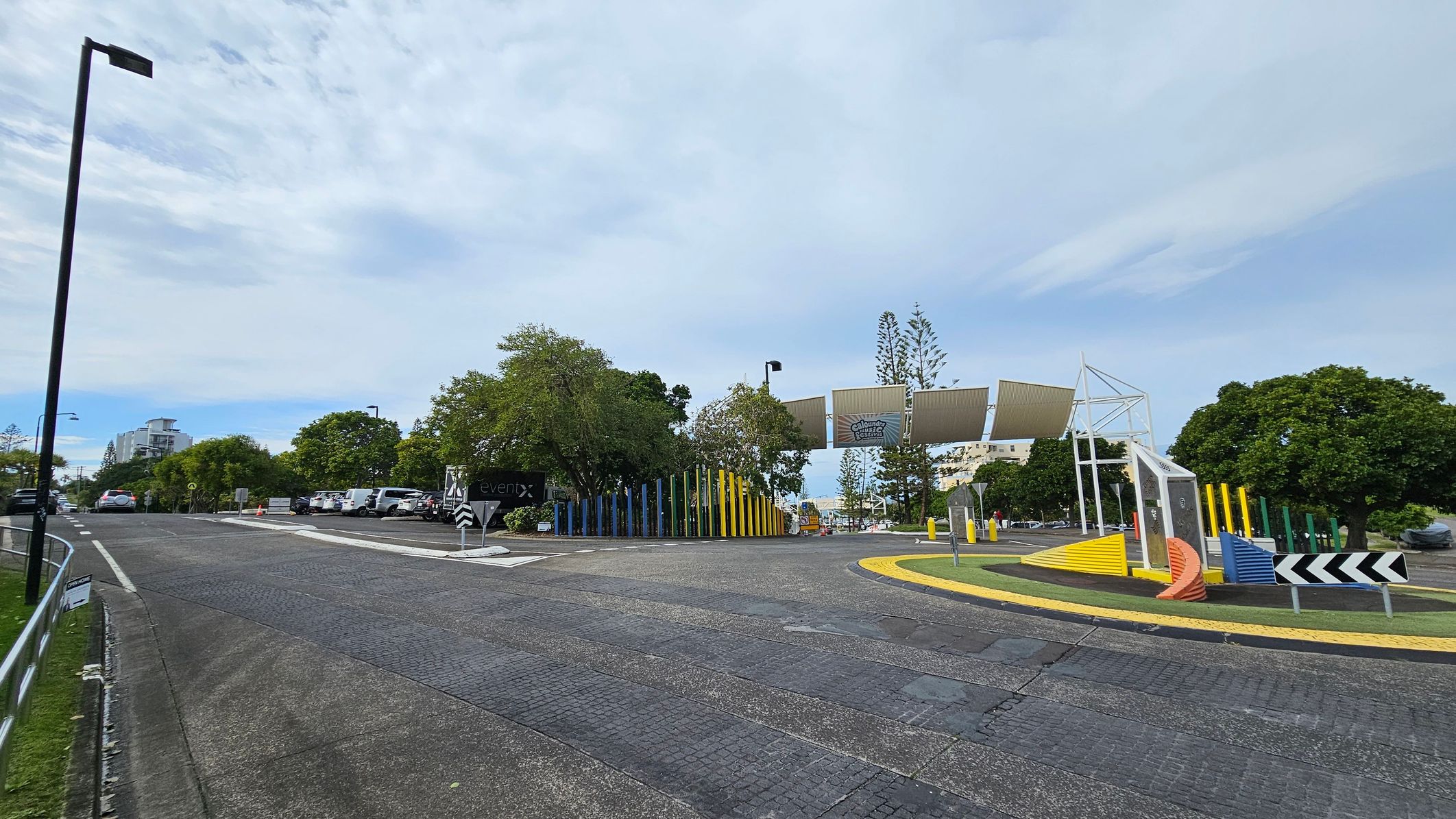 A road with a gentle curve leads to a parking area on the left, where several cars are parked. A tall streetlamp stands on the left side of the road. On the right, there is a colourful installation with vertical poles in various colours and a structure with angled panels. Trees are scattered throughout the area, and a building is visible in the distance. The road surface is textured, and there are directional signs near the installation.