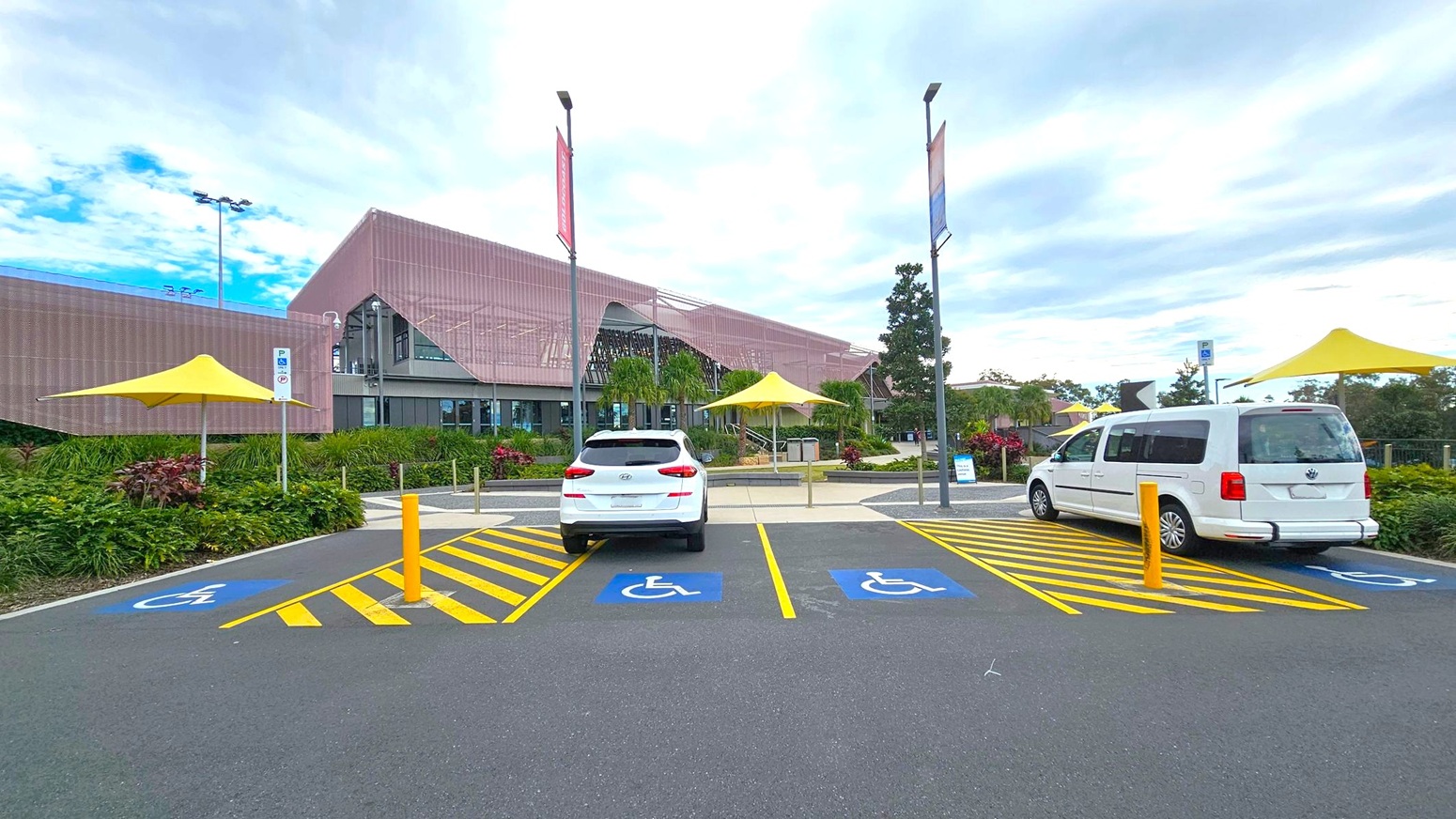 A modern building with a pink facade is in the background, featuring large glass windows and surrounded by greenery. In the foreground, there are two parked white vehicles in designated disabled parking spaces, marked with blue and yellow lines. Yellow umbrellas provide shade over nearby seating areas. Tall streetlights and banners line the pathway leading to the entrance. The ground is paved with asphalt.