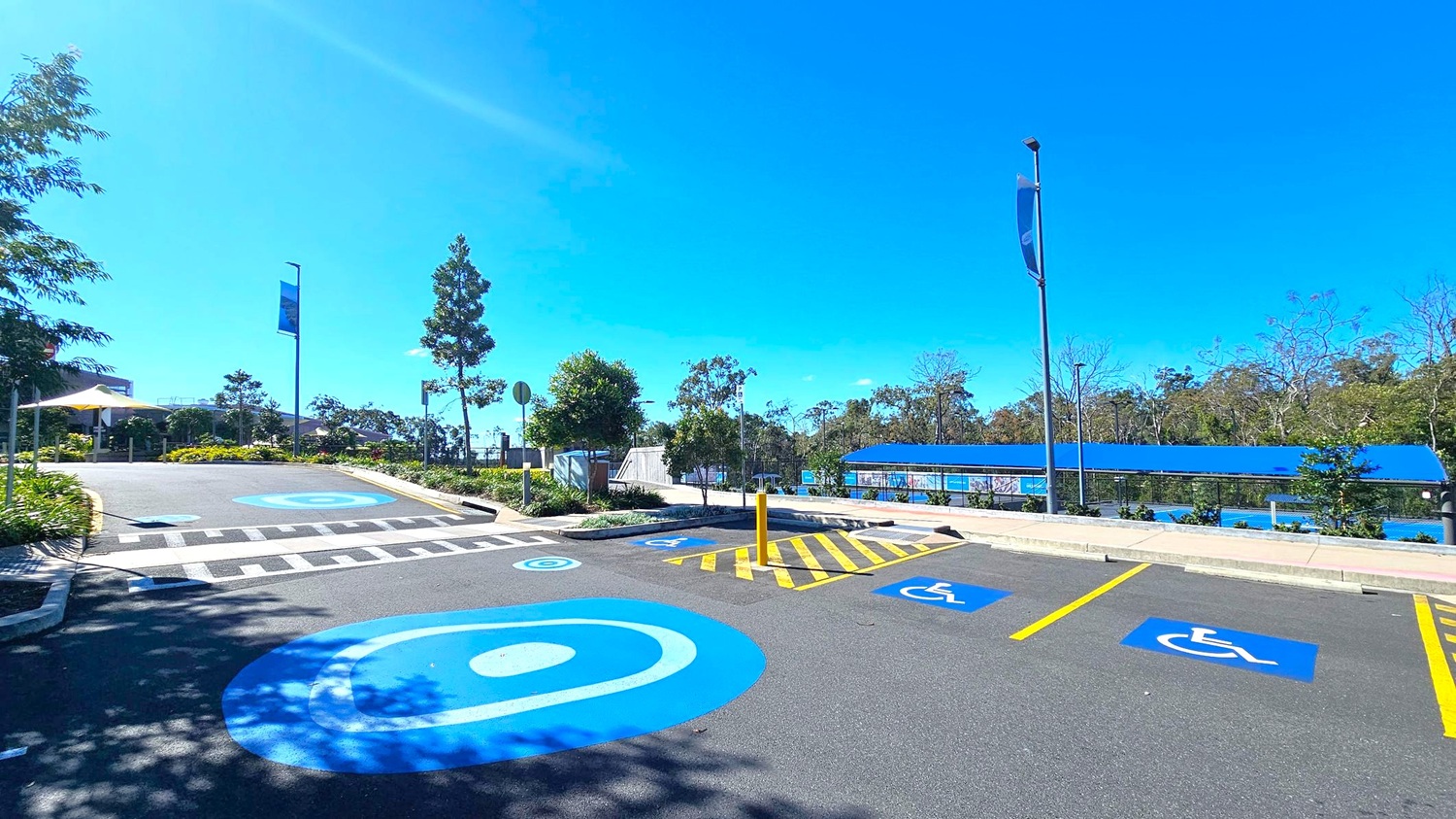 A parking area with designated disabled parking spaces marked in blue on the asphalt. On the left, there are trees and a pathway leading to a building with a white canopy. In the centre, a pedestrian crossing with white stripes is visible. On the right, a blue-roofed structure runs parallel to the parking spaces, with more trees and greenery in the background. Two tall street lamps with banners are positioned on either side of the parking area.