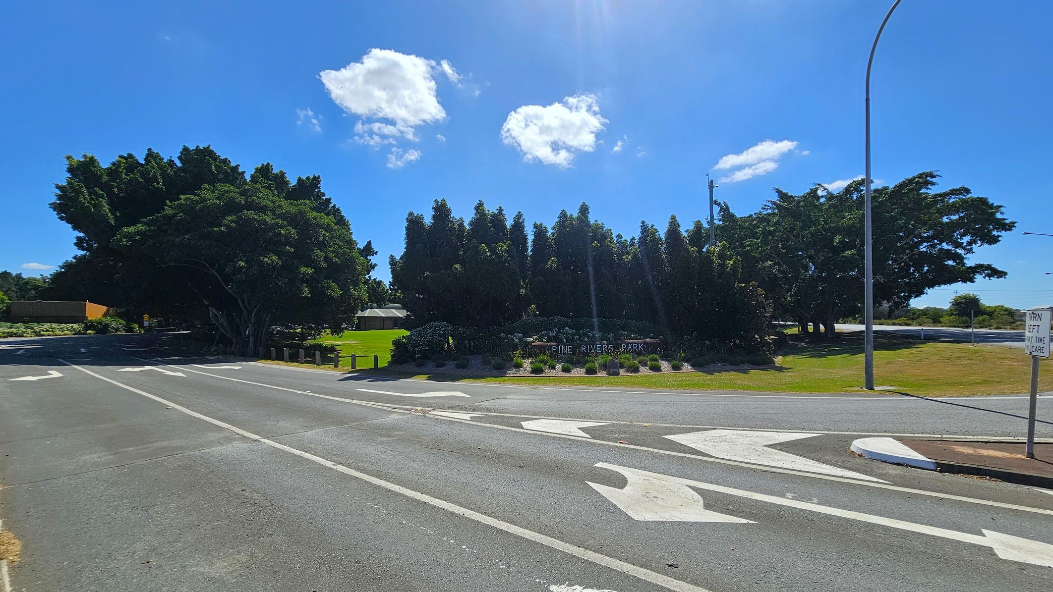 A road with white directional arrows painted on the surface. On the left, there are large, dense trees and a small building partially visible. In the centre, a sign reads "Pine Rivers Park" surrounded by neatly arranged bushes and flowers. On the right, a grassy area with a tall streetlight and a sign that says "Turn left any time with care." The sky is clear with a few clouds.