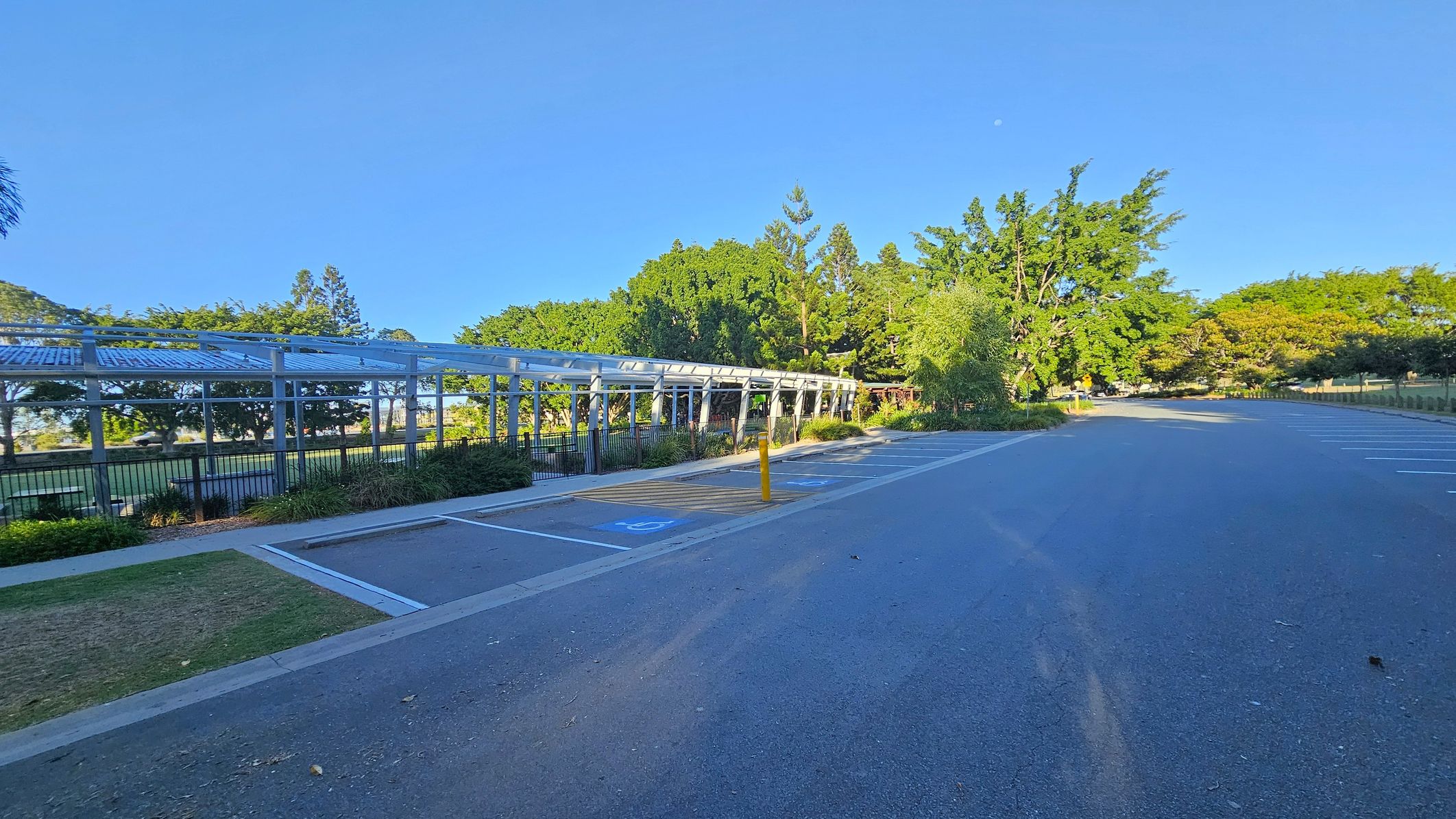 A paved parking area with marked spaces, including accessible parking spots, is on the right. A long, open-sided shelter with a metal roof runs parallel to the parking area on the left, adjacent to a grassy area with trees and shrubs. In the background, dense green trees line the area, providing a natural backdrop. A yellow bollard is visible near the centre, and the ground is mostly asphalt with some grass patches.