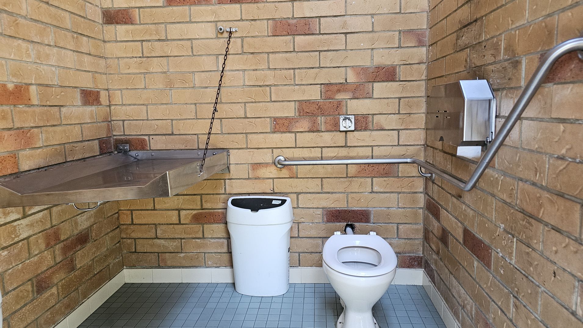 A public restroom with brick walls. On the left, a metal changing table is attached to the wall with a chain. In the centre, a white bin is placed next to a toilet. On the right, a metal handrail runs along the wall, with a toilet paper dispenser mounted above it. The floor is covered with small grey tiles.
