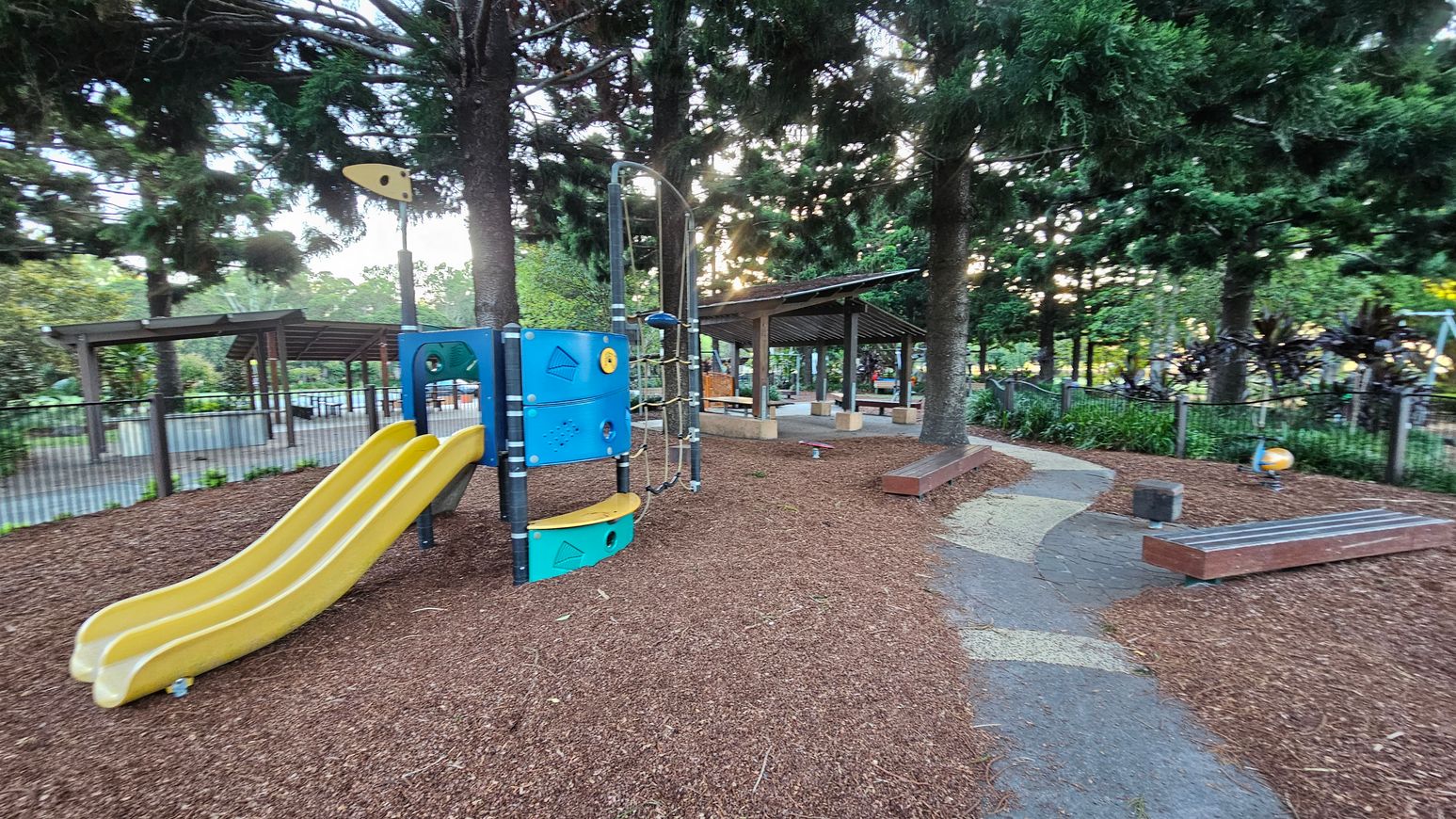 A playground with a yellow double slide on the left, attached to a blue climbing structure. The ground is covered with wood chips. In the middle, there are tall trees providing shade. To the right, a winding path leads to a sheltered picnic area with benches and tables. The background features a fence and more greenery, with additional playground equipment visible.