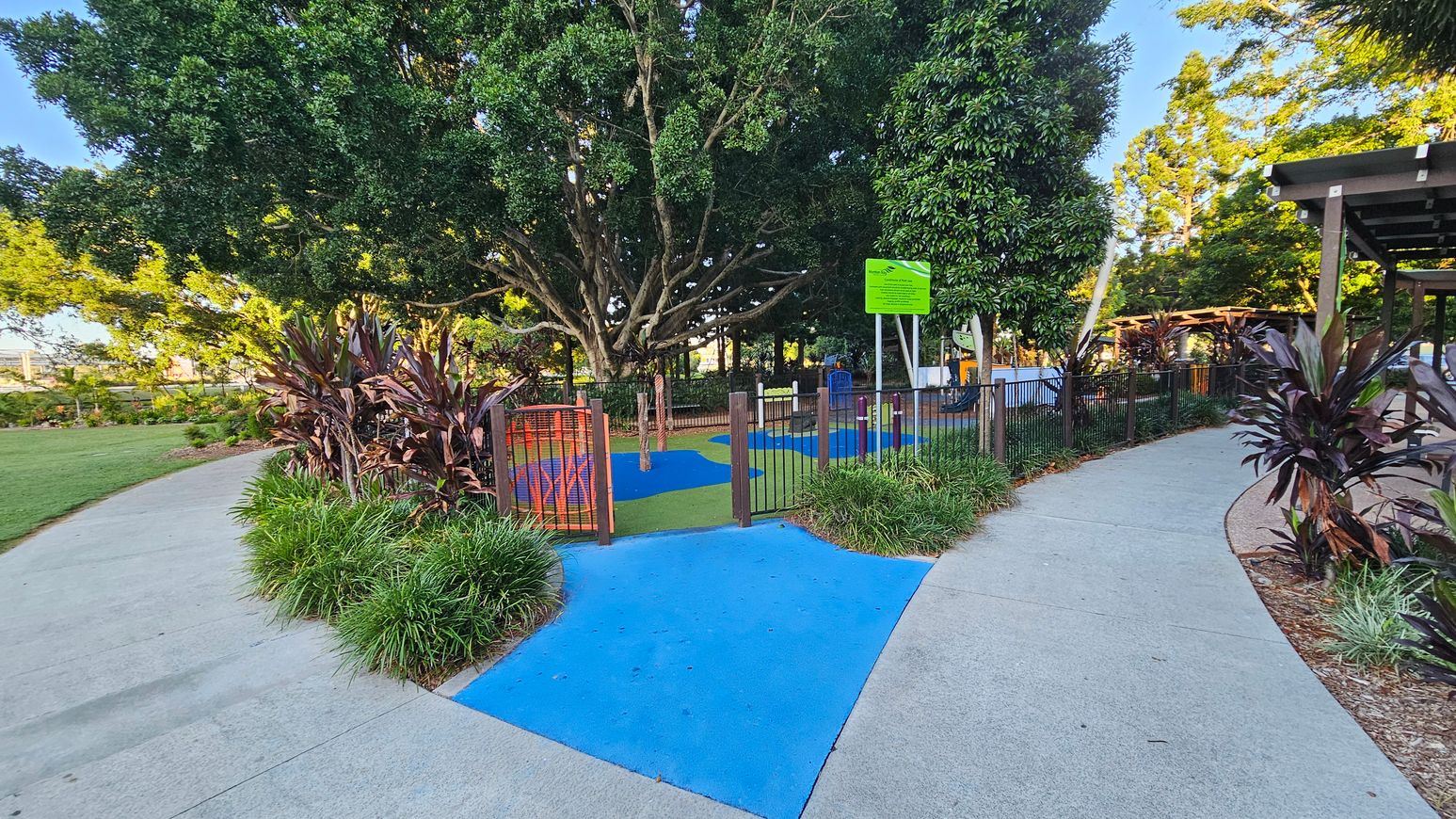 A park scene with a pathway splitting into two directions. On the left, there's a grassy area with plants and a large tree providing shade. In the centre, a fenced playground area with colourful blue and green flooring is visible, along with a sign. On the right, more plants line the path, leading to a wooden pergola structure. The ground is a mix of concrete and blue painted sections, and the area is surrounded by lush greenery.