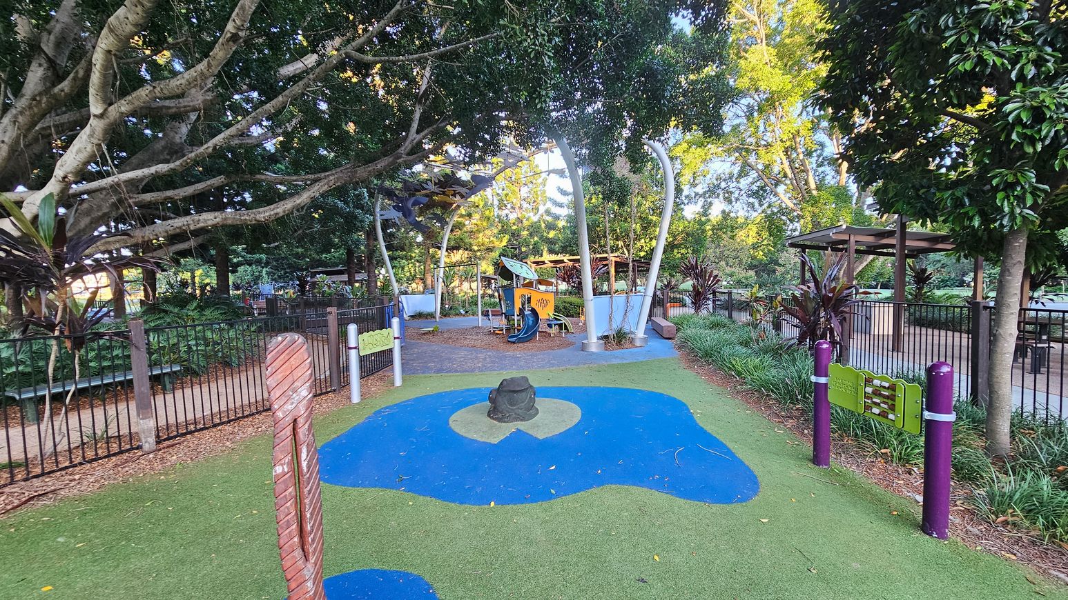 A children's playground with a soft, colourful play surface featuring green and blue areas. In the centre, there's a small sculpture resembling a frog. To the right, a musical play panel is attached to purple posts. In the background, a play structure with a slide is shaded by large trees. A fence runs along the left side, with dense foliage and a path visible beyond it. A wooden pergola is on the right, surrounded by plants and trees.
